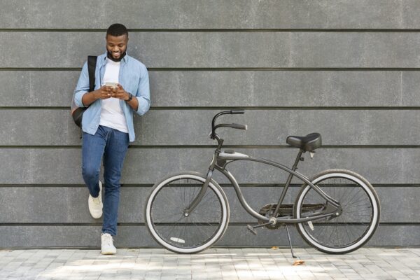 Young Black Man Using Smartphone, Standing Next To His Bike Outdoors